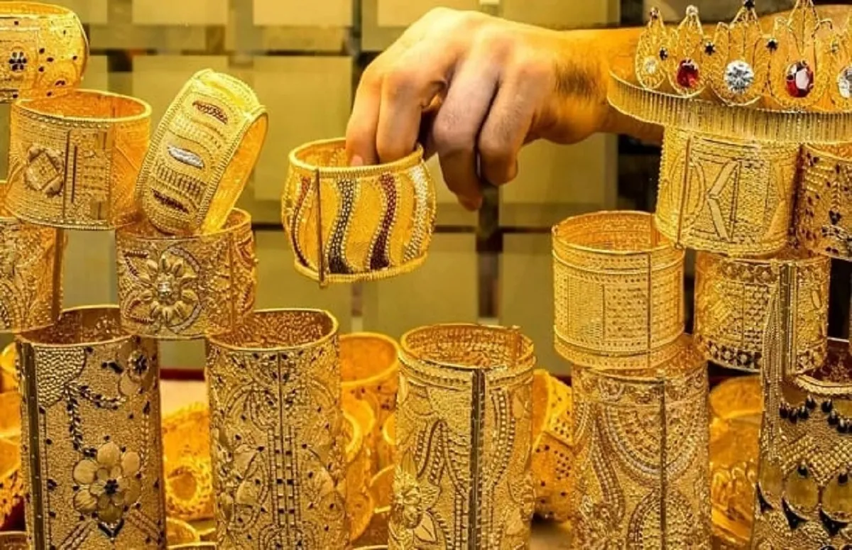 صورة عريضة لسبائك وعملات ذهبية على سطح عاكس، مع خلفية تظهر رسوم بيانية مالية ومؤشرات الأسواق، تعكس قيمة الذهب كاستثمار آمن.